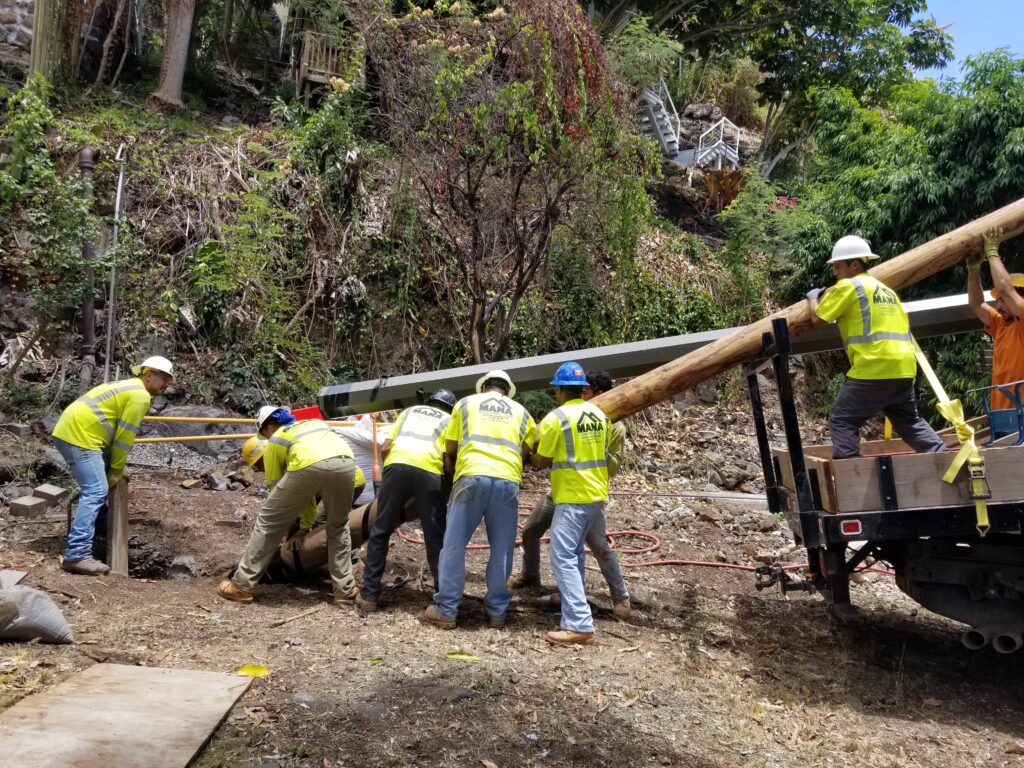 Mana crew carrying a utility pole across rough, overgrown ground